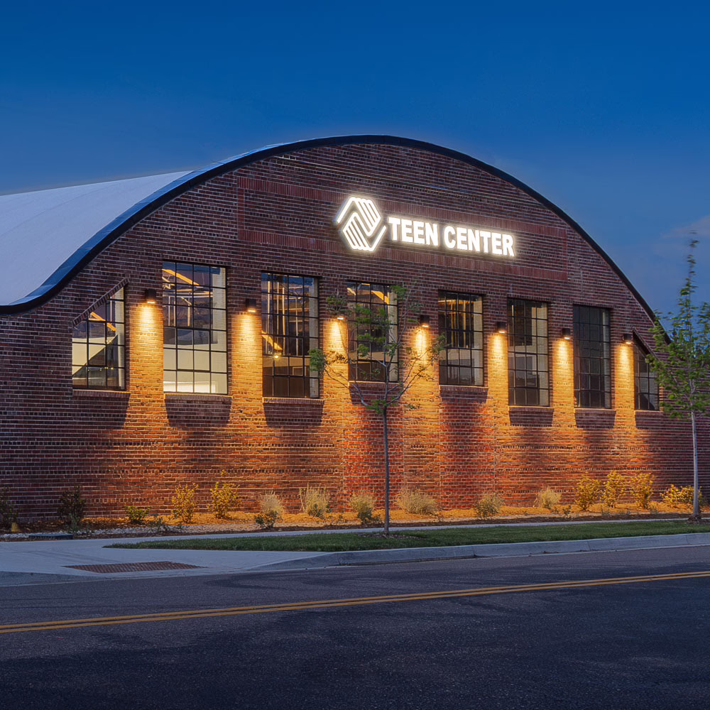 A brick building with large windows and a curved roof is lit up at dusk. A bright sign reads "Teen Center" above the entrance.