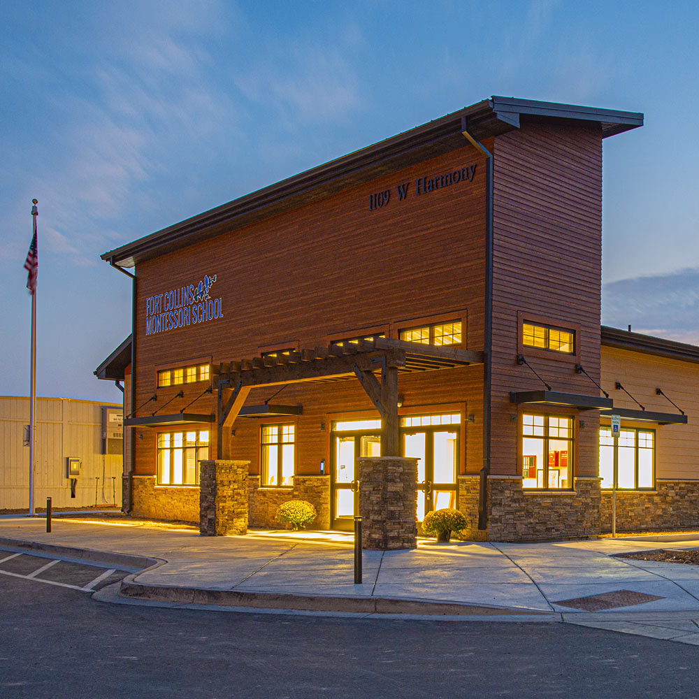 A modern building at dusk labeled "Fort Collins Montessori School" with bright windows, wooden beams, stone accents, and an American flag outside. The sky is blue with faint clouds.