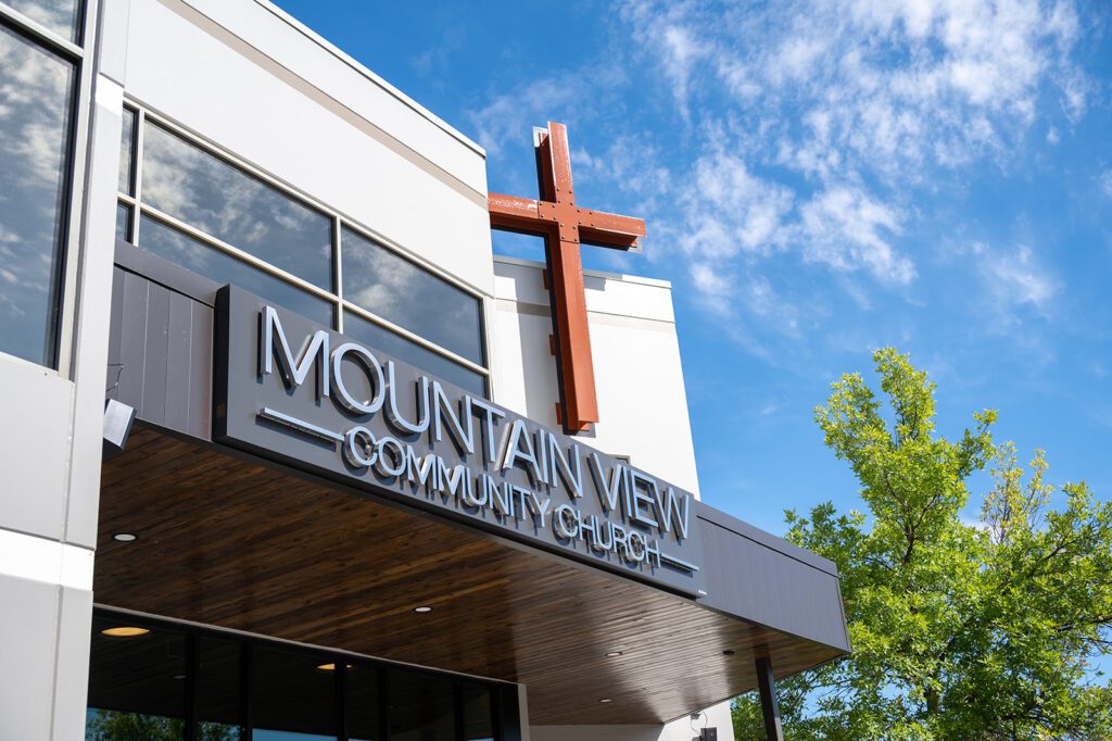 Exterior of "Mountain View Community Church" building with a large red cross displayed above the entrance. The structure has modern architecture with large windows, and the vibrant blue sky and green tree add to the welcoming atmosphere.