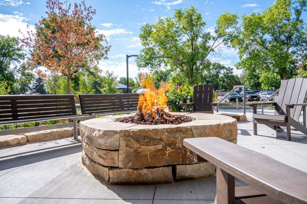A circular stone fire pit with a fire burning at its center is surrounded by wooden benches and chairs. The setting is outdoors, with green trees and a clear blue sky visible in the background. A parking lot is partially visible in the distance.