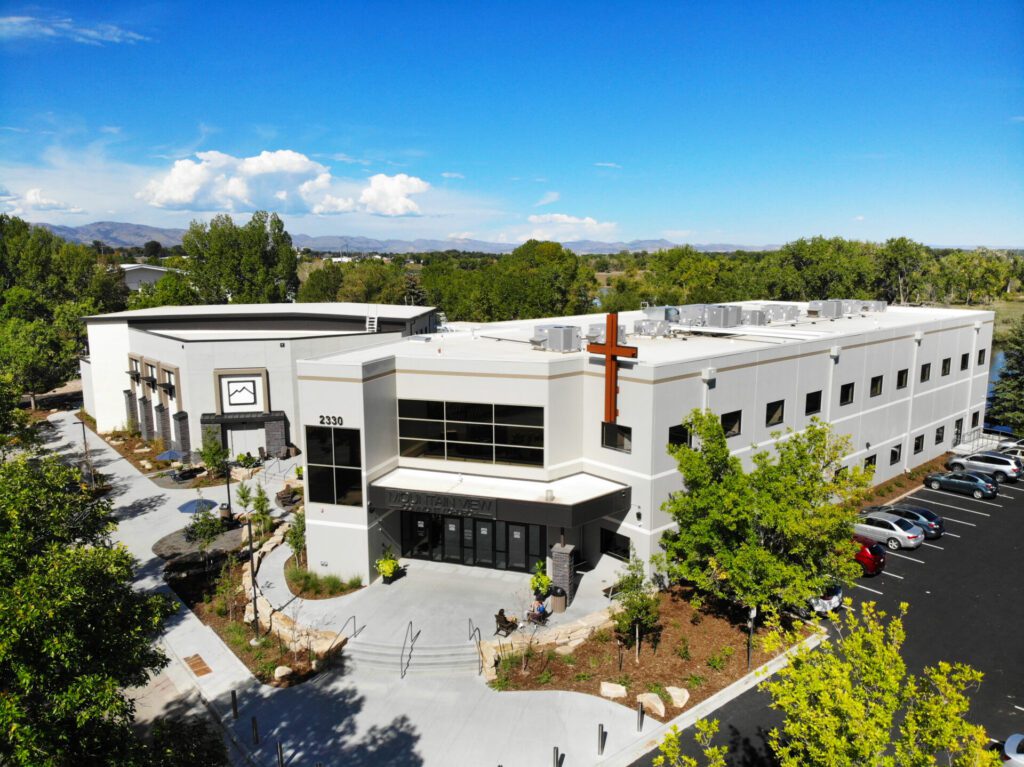 An aerial view of a modern church building with a white facade, large windows, and a prominent cross on the front. The building is surrounded by green trees and a parking lot with several cars. In the background, there are distant mountains beneath a blue sky.