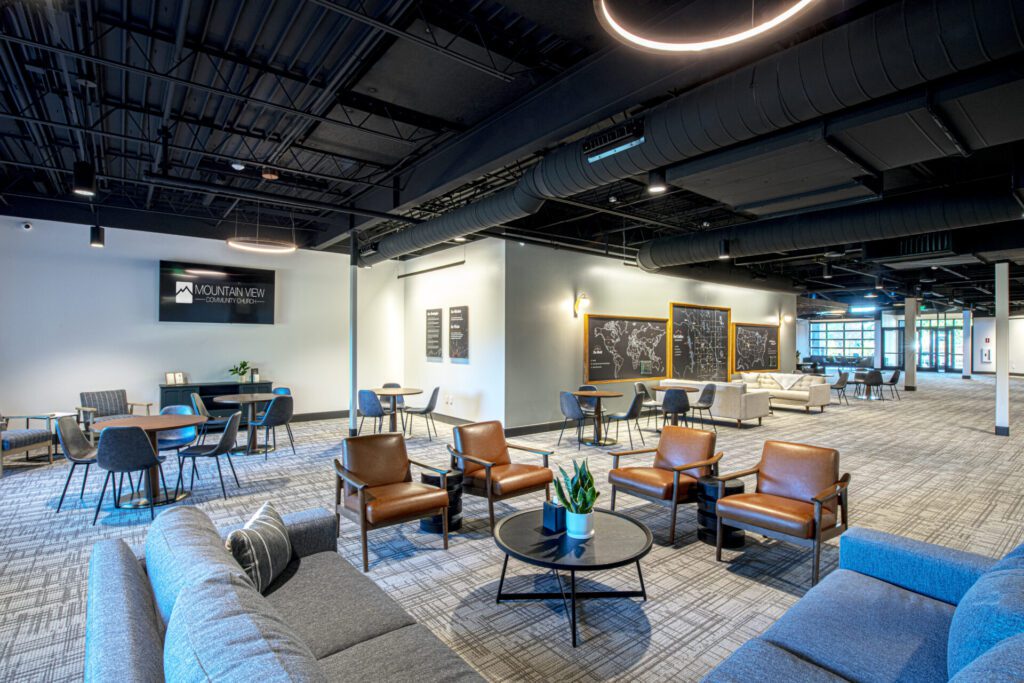 A modern lounge area with various seating options including gray sofas, wooden chairs with cushions, and round tables. The walls feature blackboards with writings, and a TV displays a logo. The space is well-lit with circular ceiling lights and large windows.