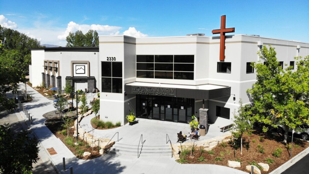 A modern church building with a large cross, labeled "Mountain View Church" at the entrance. The exterior features beige walls, large windows, and stone accents. Surrounding landscaping includes trees and shrubs. The street number 2330 is visible on the facade.