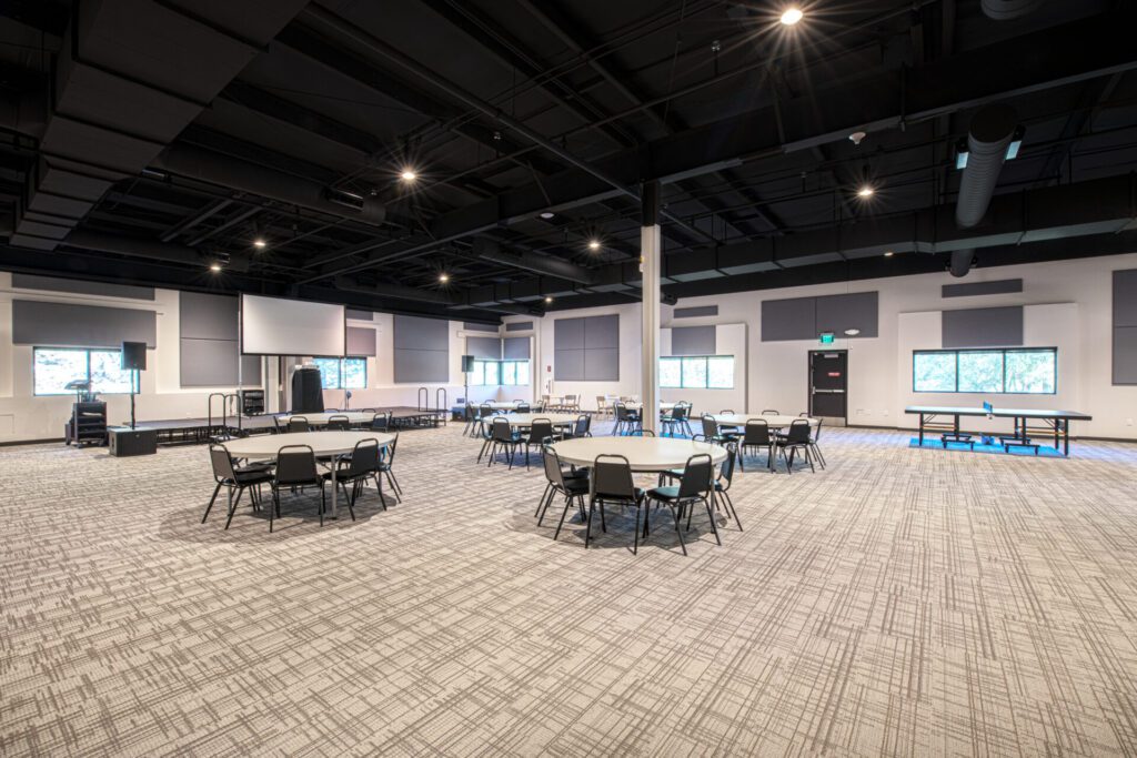 A large, modern conference room with round tables and chairs arranged throughout the space. The room features carpeted floors, high ceilings with exposed fixtures, and multiple windows allowing natural light. A stage is present in the far corner of the room.