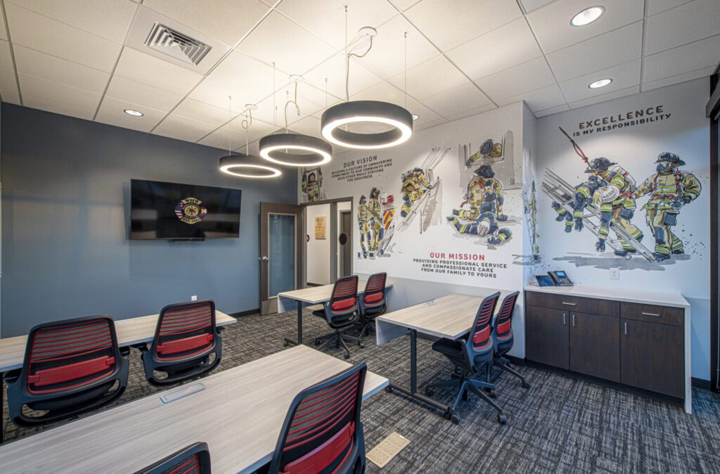 A modern training room with a large wall mural depicting firefighters in action. The room has several workstations with chairs, a mounted TV screen, and ceiling lights. Motivational text on the walls emphasizes the mission and responsibility of the fire department.