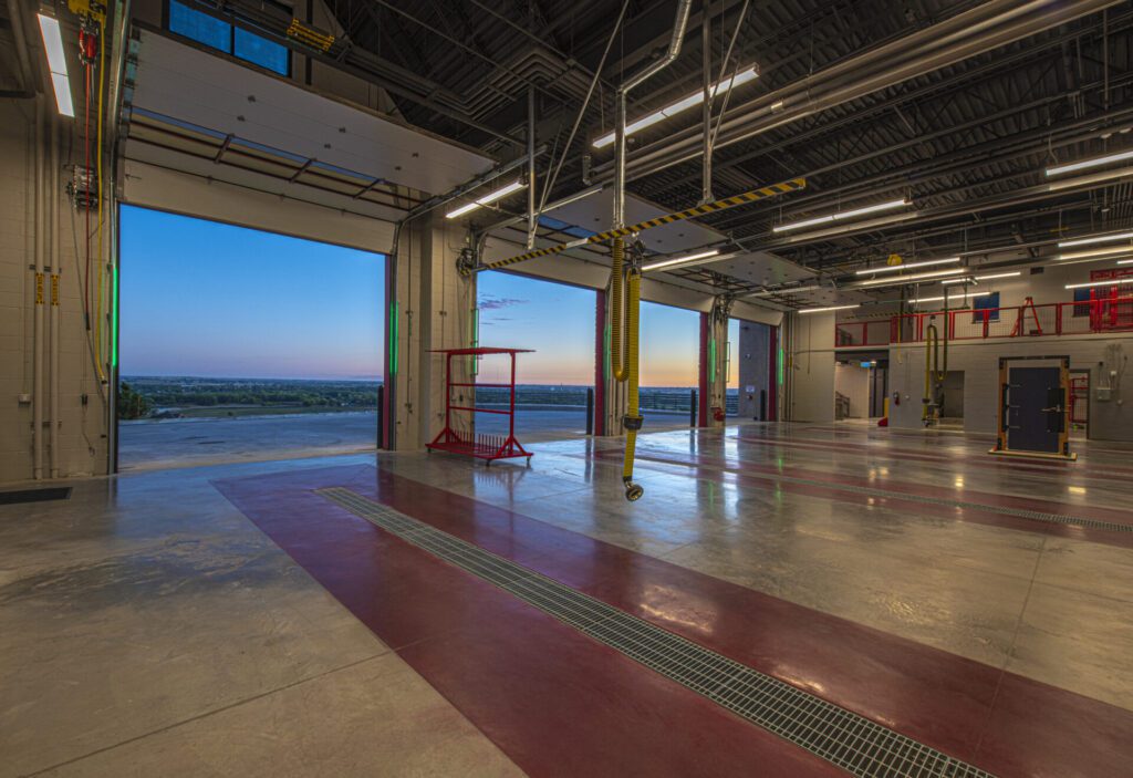 An empty, spacious industrial garage with three large open doors facing a scenic evening landscape. The well-lit interior has polished concrete floors, various hanging hoses, and ceiling pipes. The sky outside is painted with hues of blue and orange from the setting sun.