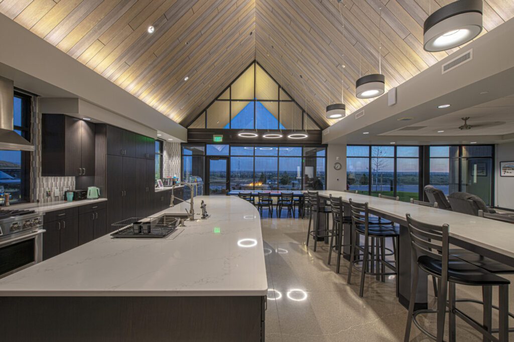 A modern, spacious kitchen with high, wooden vaulted ceilings featuring recessed lighting and hanging pendant lights. Large windows at the end of the room offer a scenic view. The kitchen includes a long island with bar stools, white countertops, and dark cabinetry.