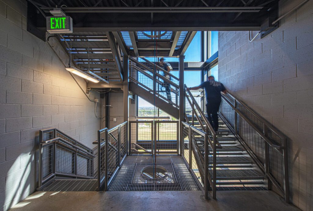 A person stands on a metal staircase in an industrial-style building next to a large window. Another person is slightly visible on the upper level. An "EXIT" sign is illuminated on the left wall. The staircase has metal railings and mesh steps.