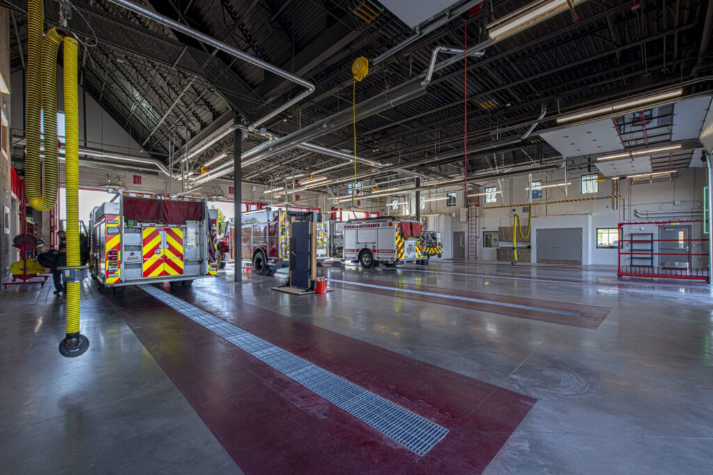 Interior of a large, modern fire station with two fire trucks parked inside. The space is clean and spacious, with high ceilings, various equipment, and bright lighting. Overhead hoses and other firefighting tools are visible throughout the facility.