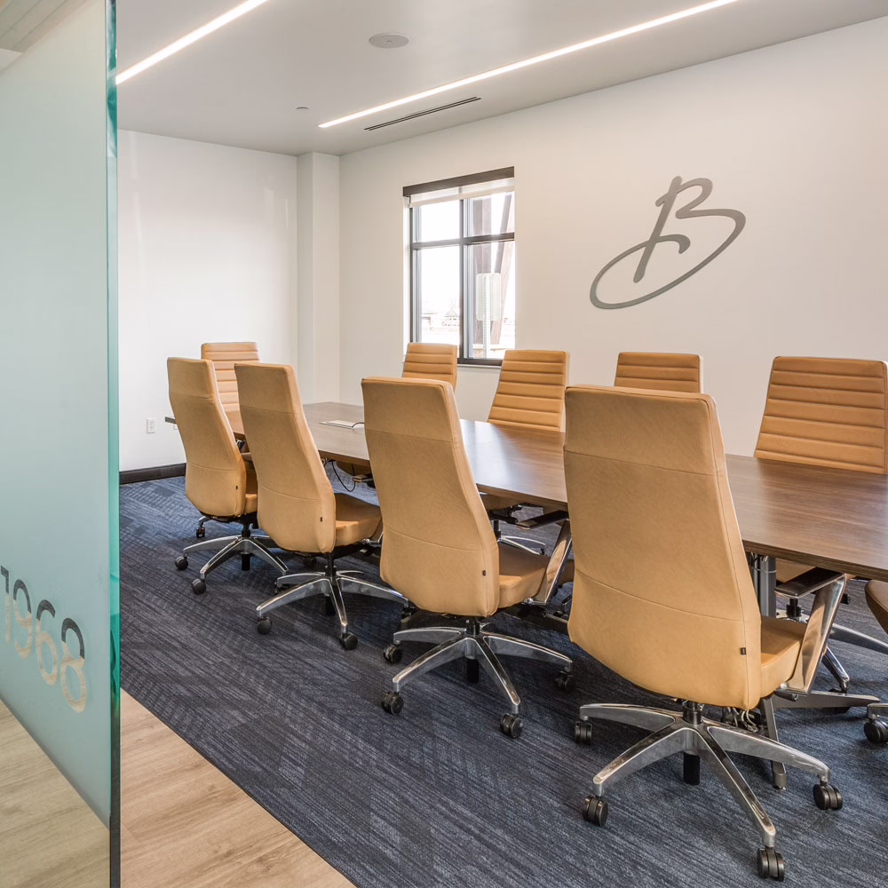 A modern conference room with a long wooden table surrounded by tan leather chairs on wheels, blue carpet, white walls, a window, and a stylized "B" logo on the wall.