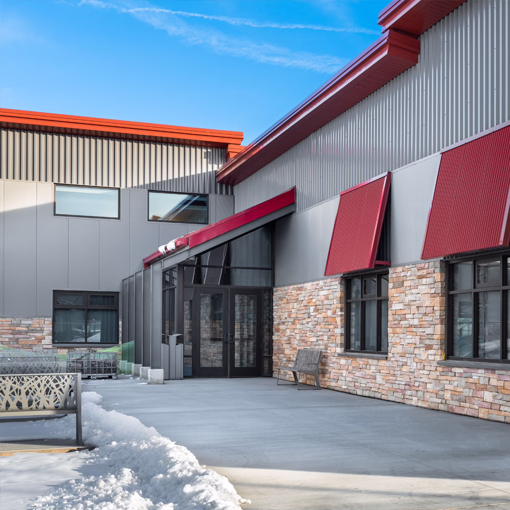 A modern building with stone and metal siding, large glass entrance doors, red awnings, and a snow-lined walkway under a blue sky. A bench sits near the entrance.