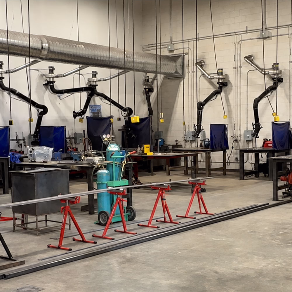 A welding workshop with multiple workstations, welding machines, black exhaust arms on the walls, red stands, and metal pipes on the floor. The area is spacious, organized, and has industrial lighting.