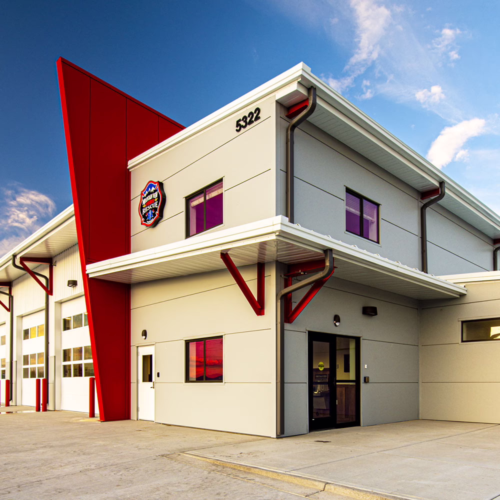 A modern fire station building with gray walls, large red structural accents, multiple garage doors, and the address 5322 displayed on the front under a blue sky.