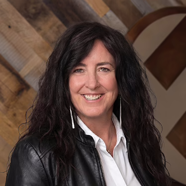 A woman with long, wavy dark hair smiles at the camera. She is wearing a black leather jacket over a white shirt, with long earrings. The background features wooden planks arranged diagonally.